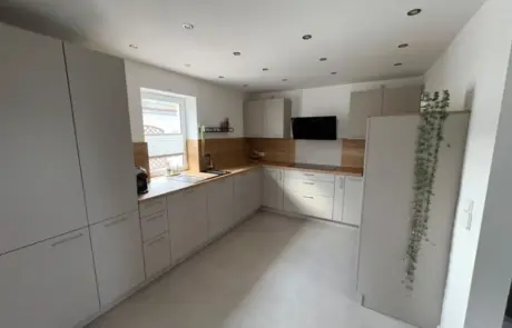 Moderne Küche mit weißen Schränken, einer Küchenrückwand aus Holz, Einbaugeräten, einem Fenster über der Spüle und einem hohen Schrank, an dessen Seite eine grüne Hängepflanze hängt.