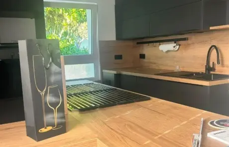 Auf einer hölzernen Kücheninsel in einer modernen Küche mit schwarzen Schränken steht eine Geschenktüte mit goldenen Weinglas-Silhouetten. Auf einer hölzernen Kücheninsel in einer modernen Küche mit schwarzen Schränken steht eine Geschenktüte mit goldenen Weinglas-Silhouetten.