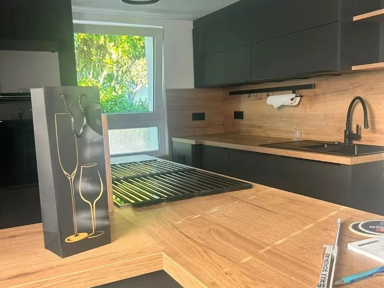 Auf einer hölzernen Kücheninsel in einer modernen Küche mit schwarzen Schränken steht eine Geschenktüte mit goldenen Weinglas-Silhouetten.